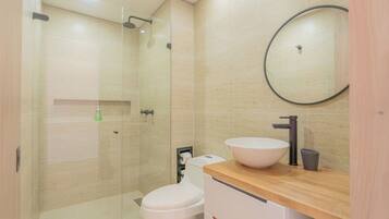 Une salle de bain moderne avec une douche à parois de verre, des toilettes blanches, un meuble-lavabo en bois doté d’un grand évier et un miroir rond.