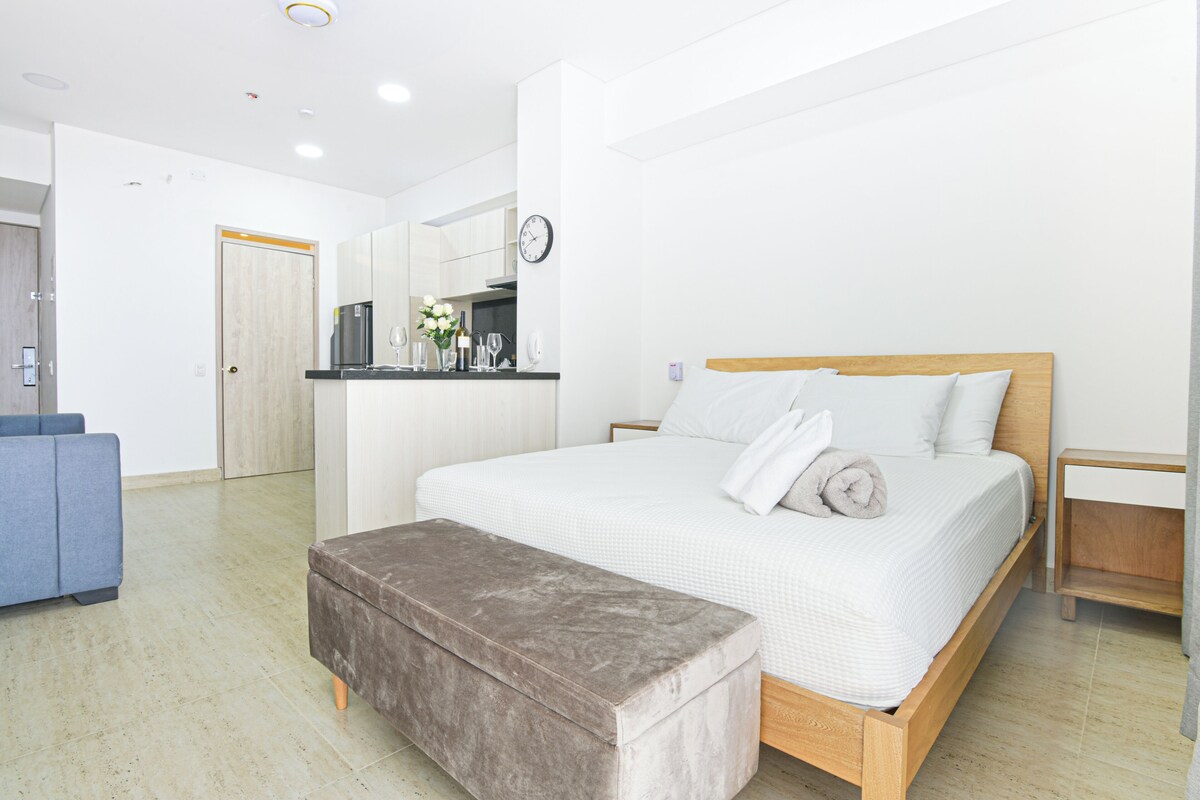A modern bedroom with a bed, bedside tables, a bench, a kitchenette, and a bathroom.