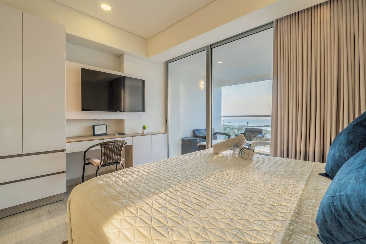A modern hotel room with a large bed, a desk with a flat-screen TV, a chair, and a view of the balcony and cityscape.