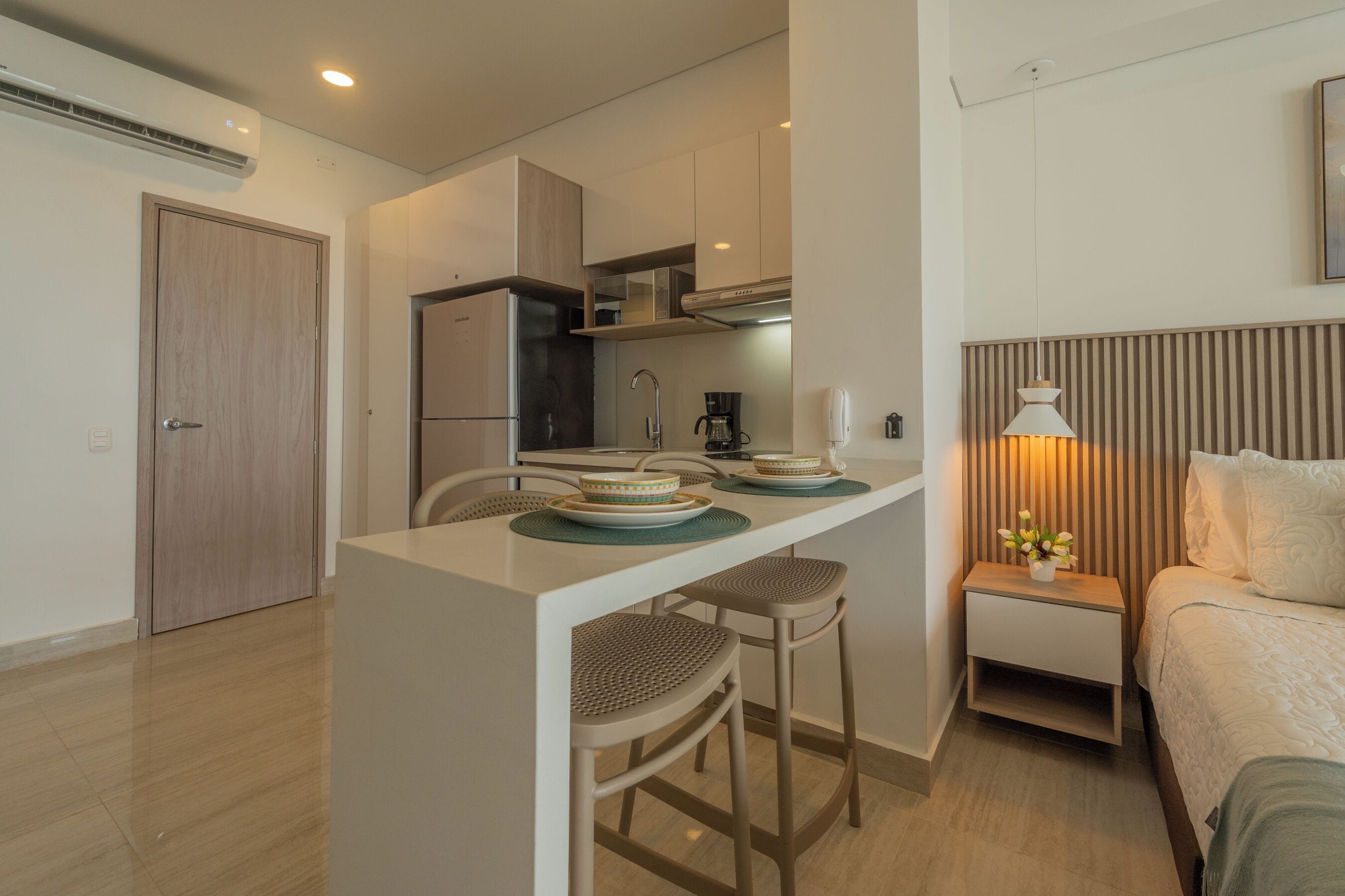 A compact kitchen with a central island, a refrigerator, and a coffee maker. There is a small bedside table with a lamp and a plant.