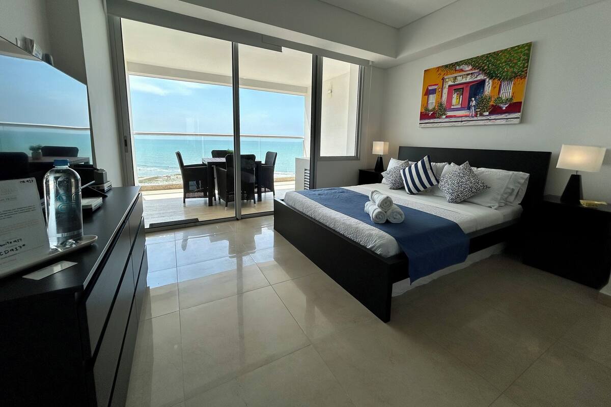 A modern hotel room with a bed, bedside tables, a desk with a water bottle, a chair, and a view of the beach and ocean through large glass doors.