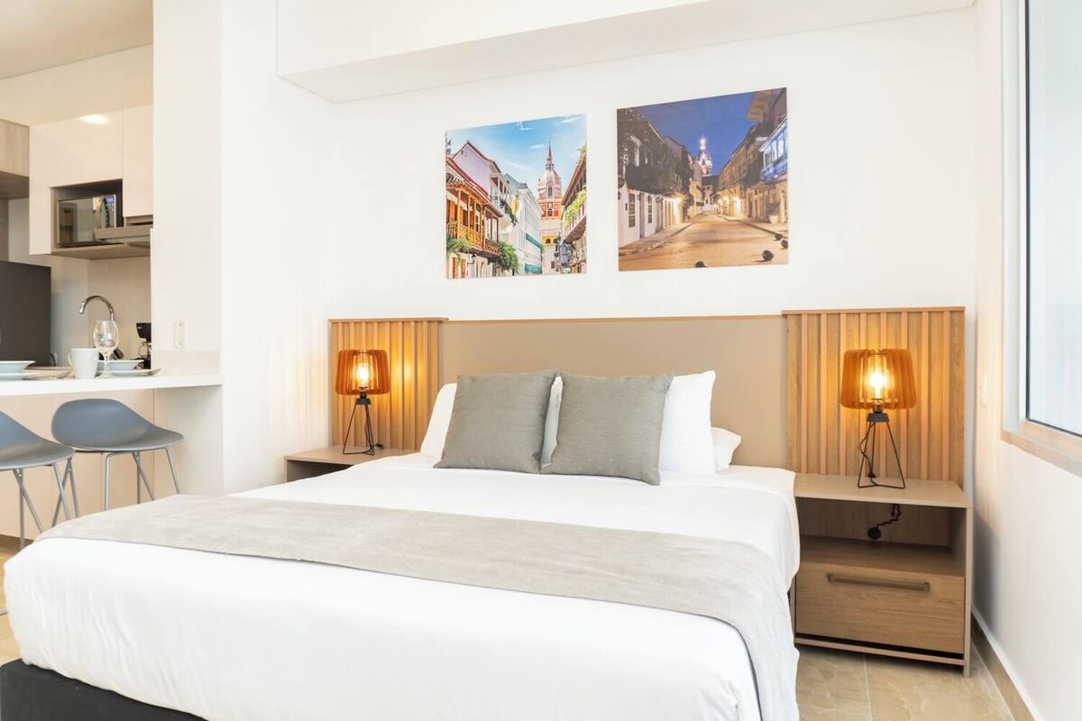 A modern hotel room with a large bed, two bedside tables with lamps, a desk with a chair, and two framed pictures on the wall.