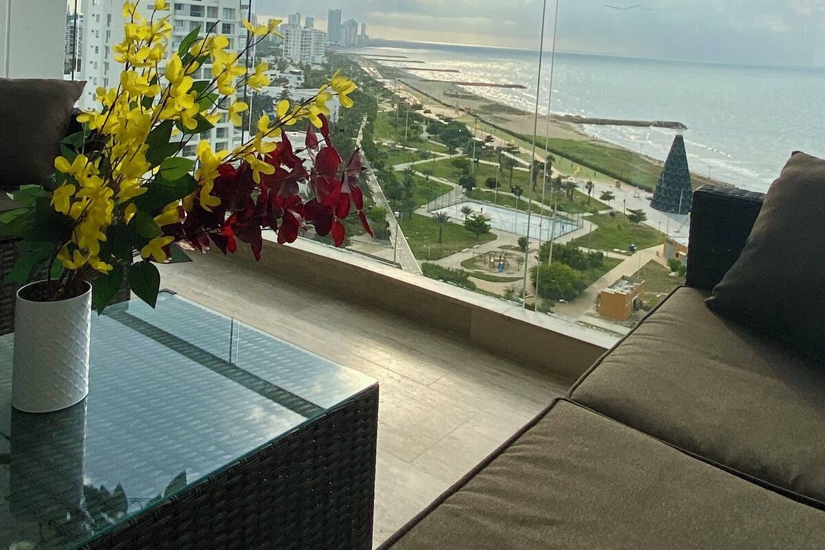 A balcony with a view of a beach and cityscape, a wicker table, and a vase with yellow flowers.