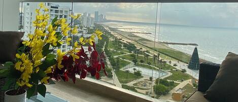 A balcony with a view of a beach and cityscape, a wicker table, and a vase with yellow flowers.