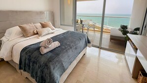 Un dormitorio moderno con una cama grande, un balcón con vistas al mar y un pequeño comedor.