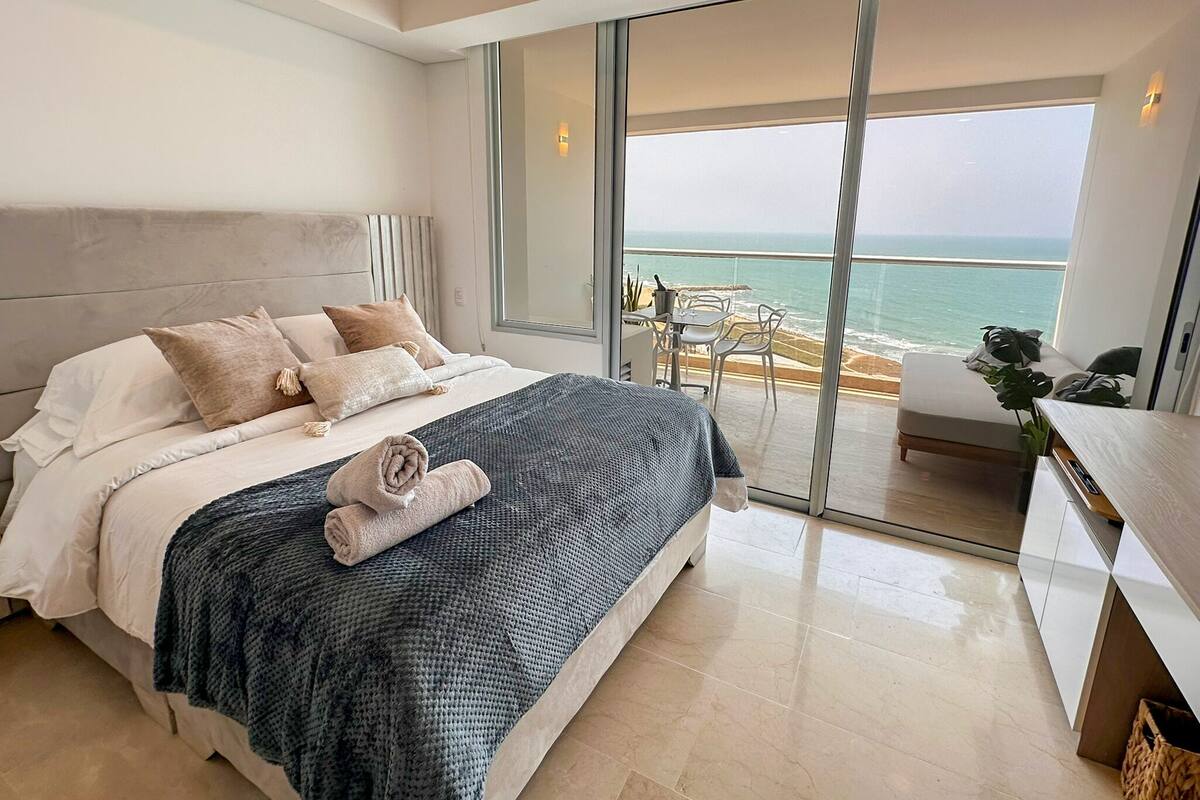 A modern bedroom with a large bed, a balcony with a view of the sea, and a small dining area.