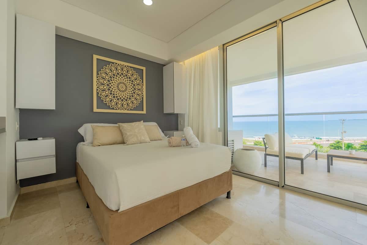 A modern bedroom with a large bed, a nightstand, a decorative wall piece, and a view of the beach through large sliding doors.