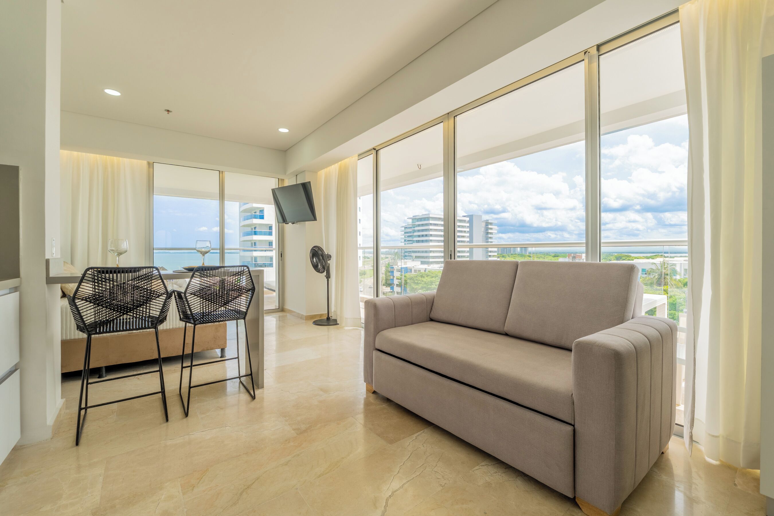 A modern living room with a sofa, two black chairs, a flat-screen TV, and a large window offering a city view.