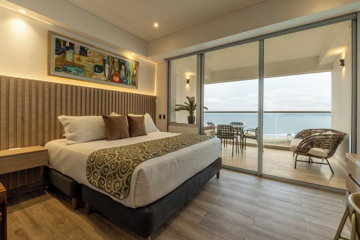 A bedroom with a large bed, a painting on the wall, and a balcony with a view of the sea.