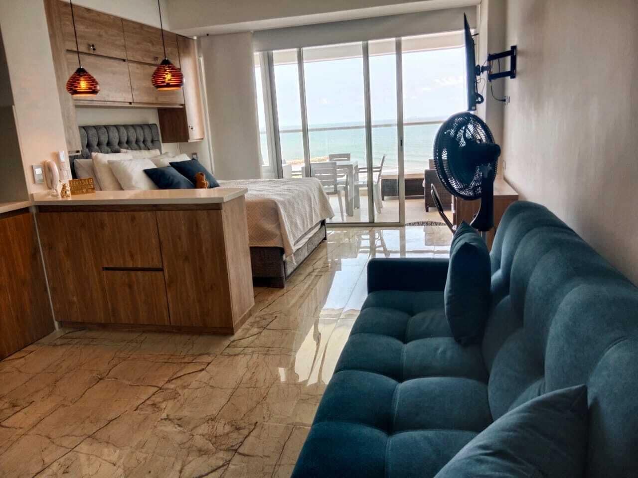A modern hotel room with a bed, a sofa, a small kitchen area, and a balcony with a view of the sea.