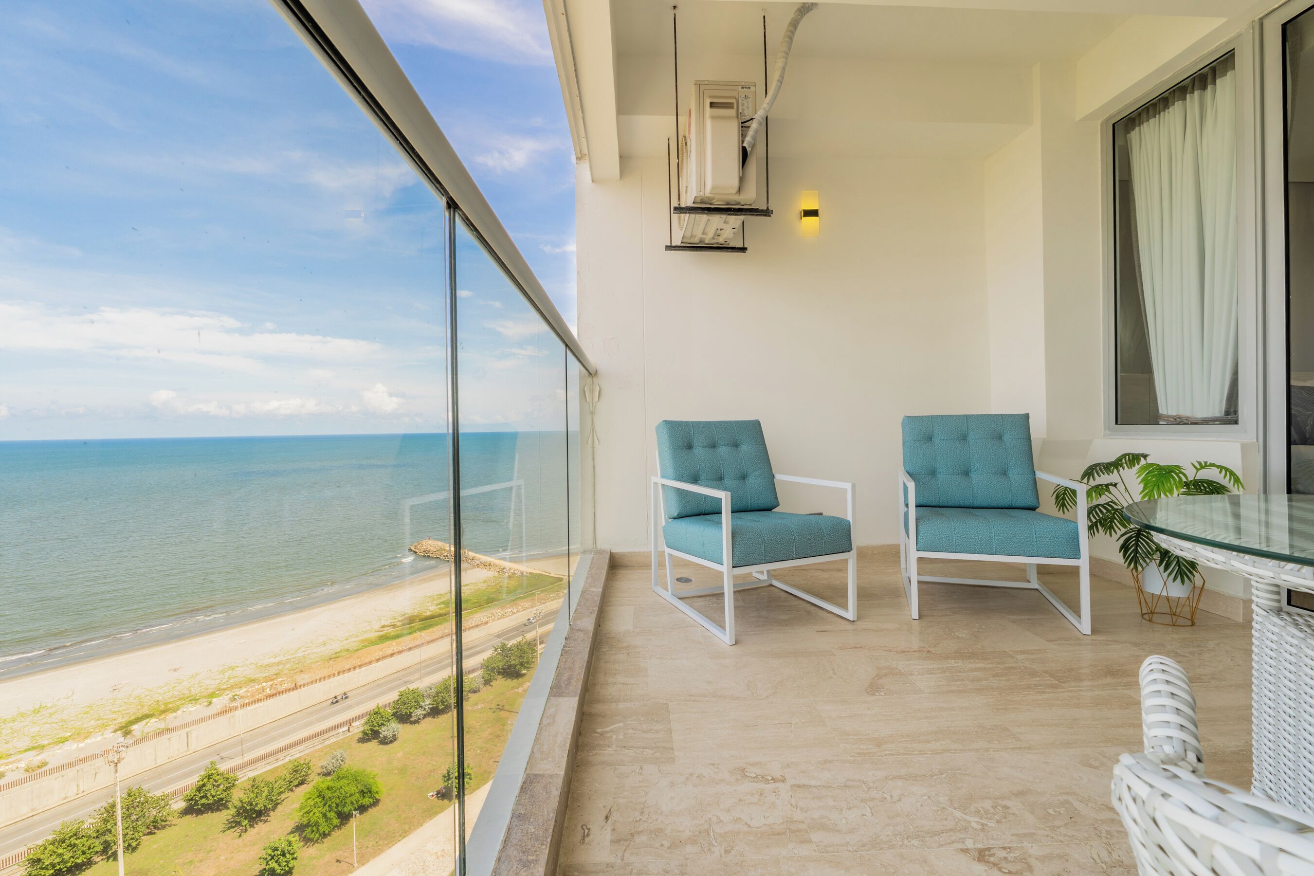 A balcony with a view of the sea, two chairs, a potted plant, and a glass table.
