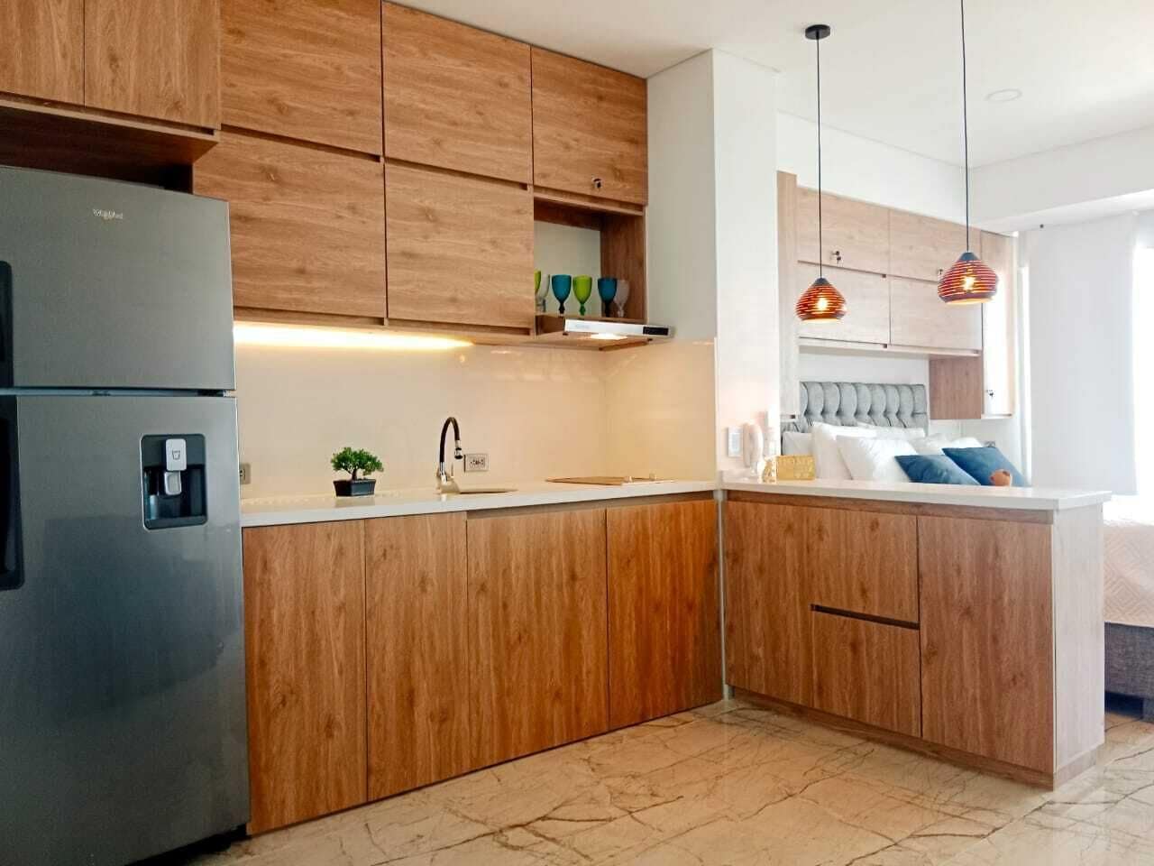 A modern kitchen with wooden cabinets, a stainless steel refrigerator, a sink, and pendant lighting.