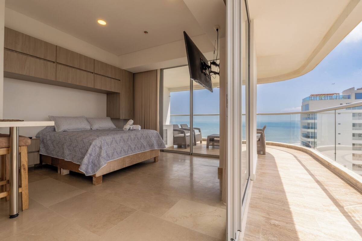 A modern bedroom with a bed, a desk, and a balcony with a view of the sea.