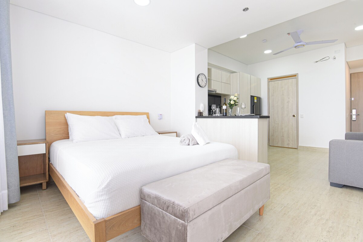A modern bedroom with a wooden bed, a bedside table, a bench, a kitchenette, and a ceiling fan.