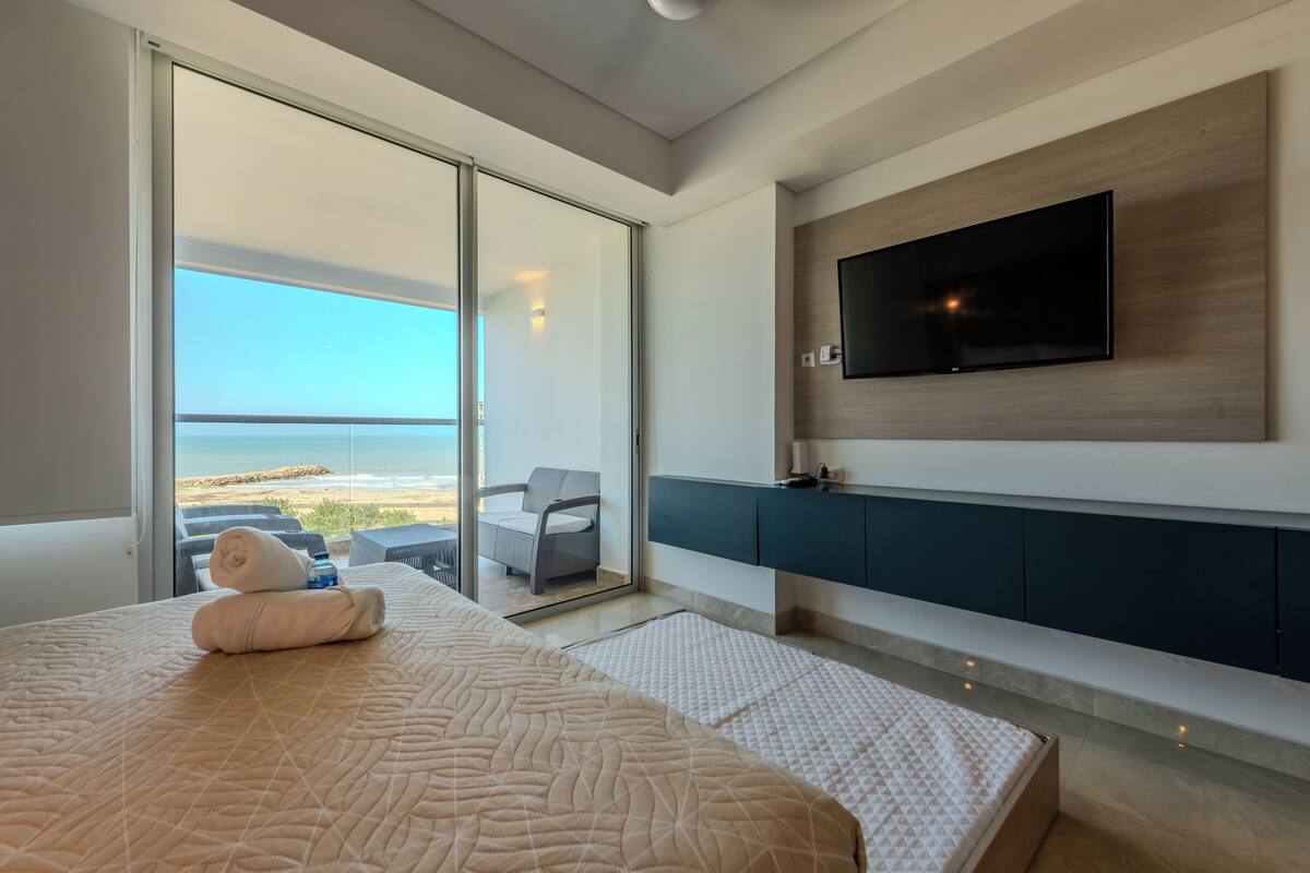 A modern hotel room with a large bed, a TV mounted on the wall, and a balcony with a view of the beach.
