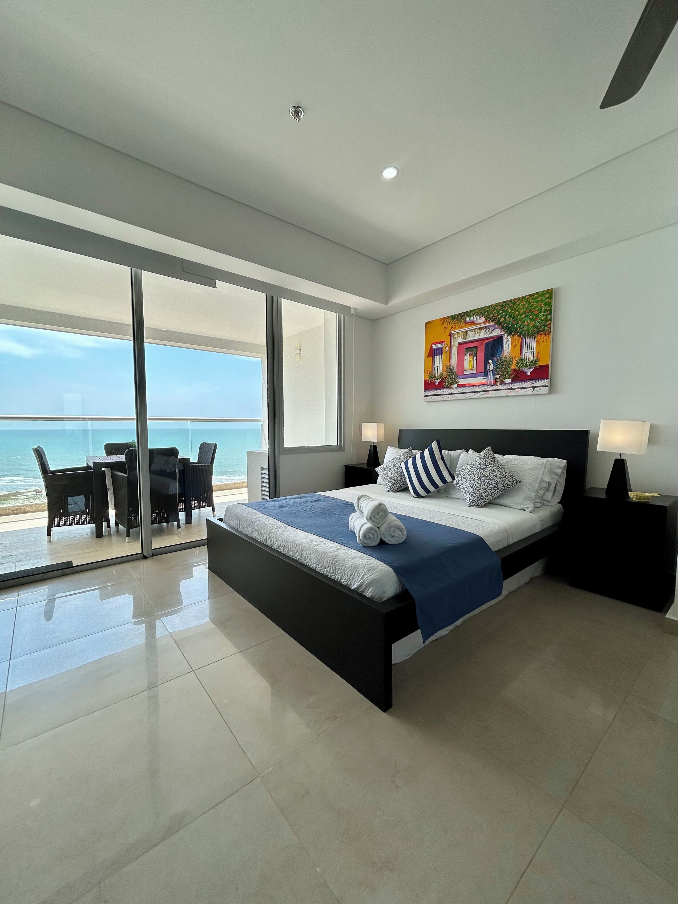 A modern bedroom with a large bed, a balcony with a view of the sea, and a painting on the wall.