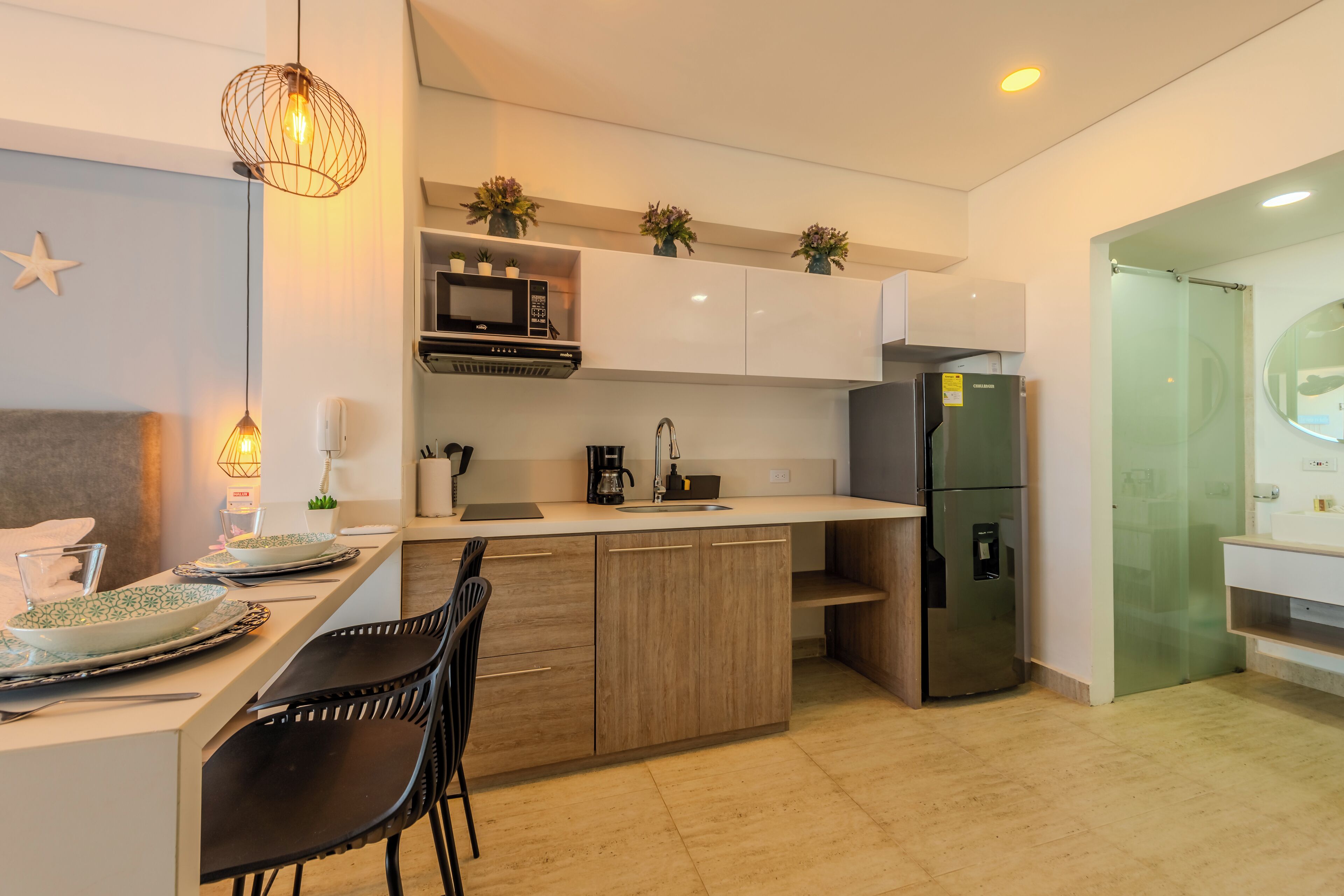 A modern kitchen with a central island, black chairs, a microwave, a refrigerator, and decorative plants.
