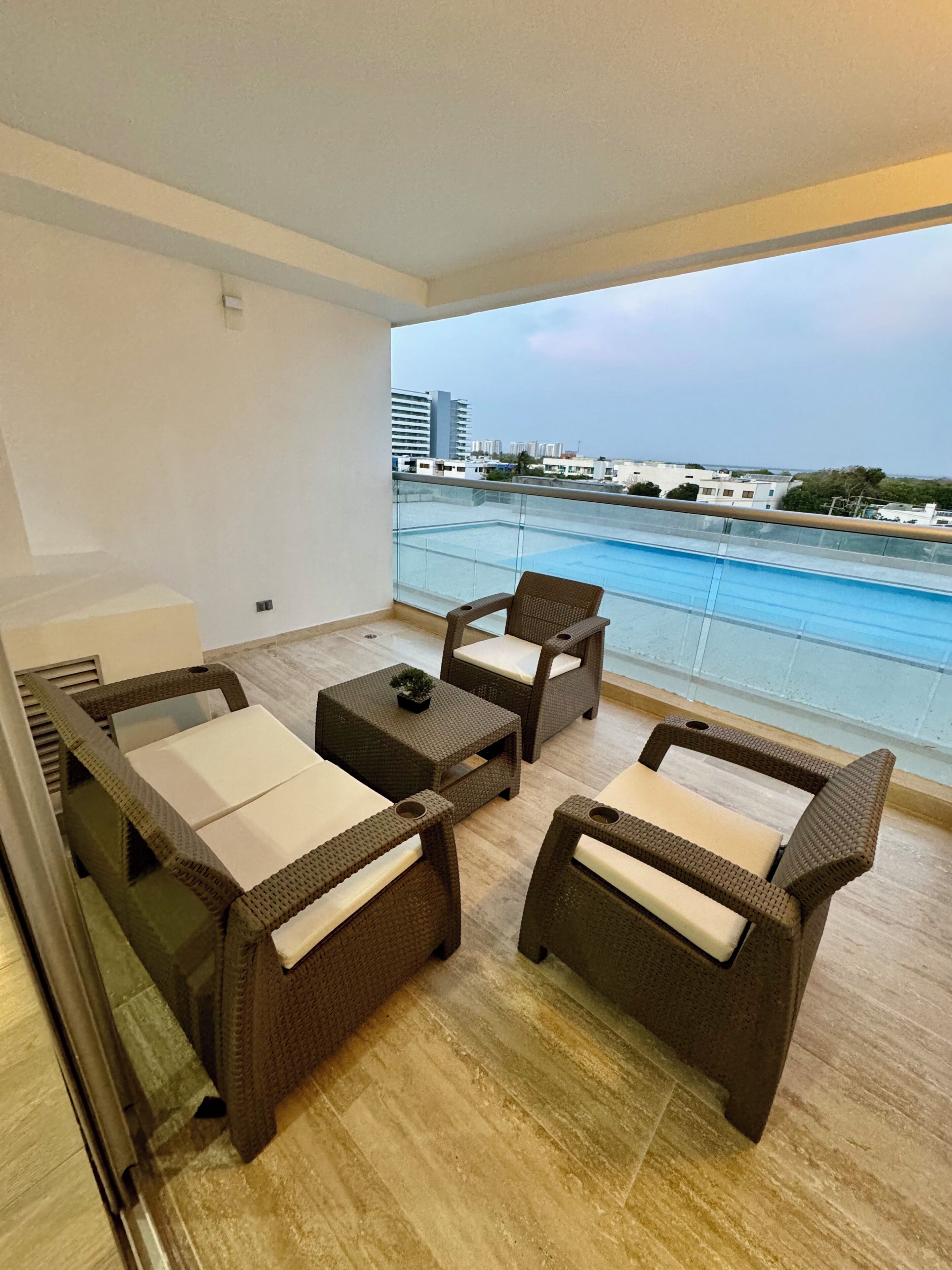 A balcony with wicker furniture, a small table, and a view of the sea and buildings.