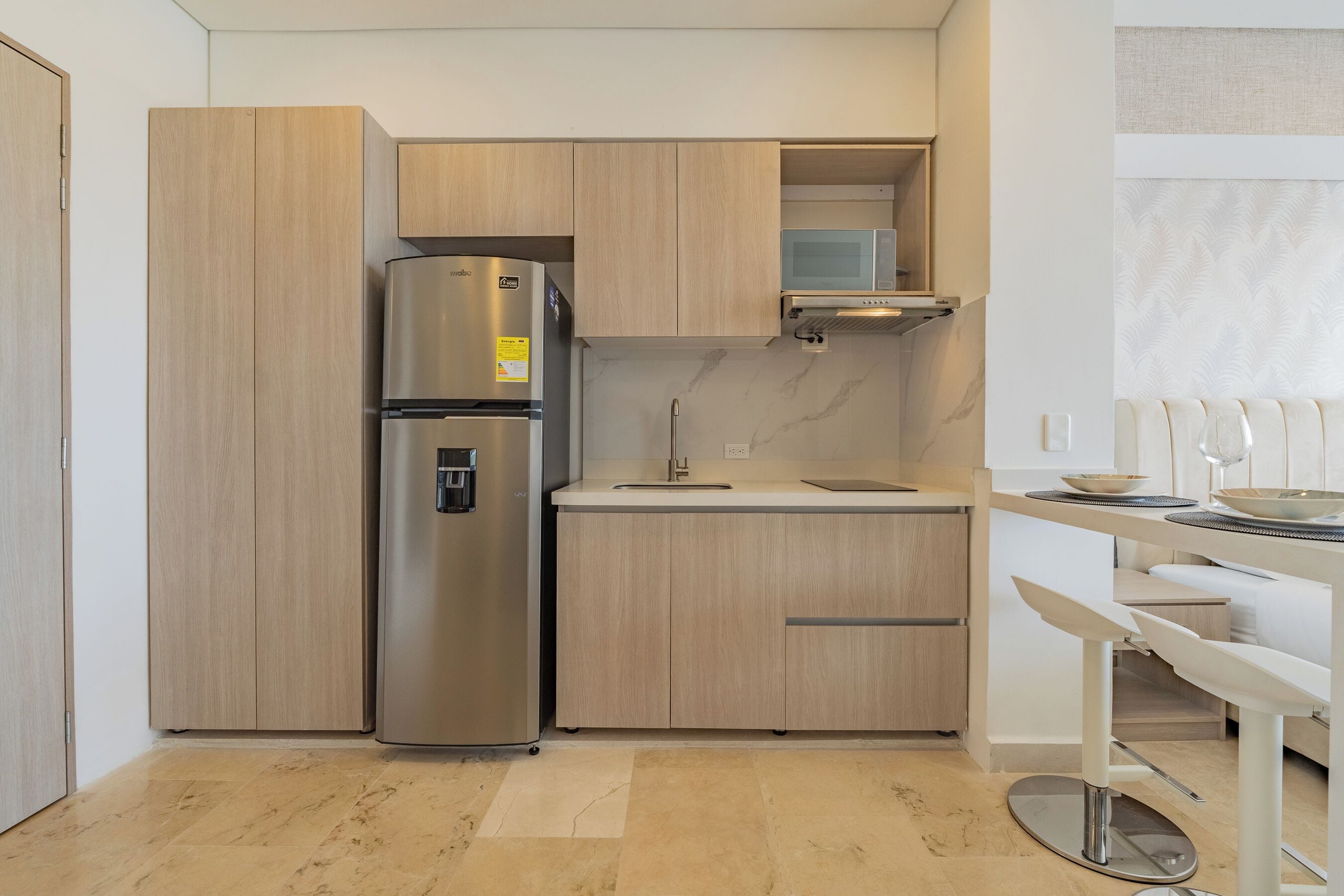 A compact kitchen with a stainless steel refrigerator, wooden cabinets, a microwave, and a white countertop.