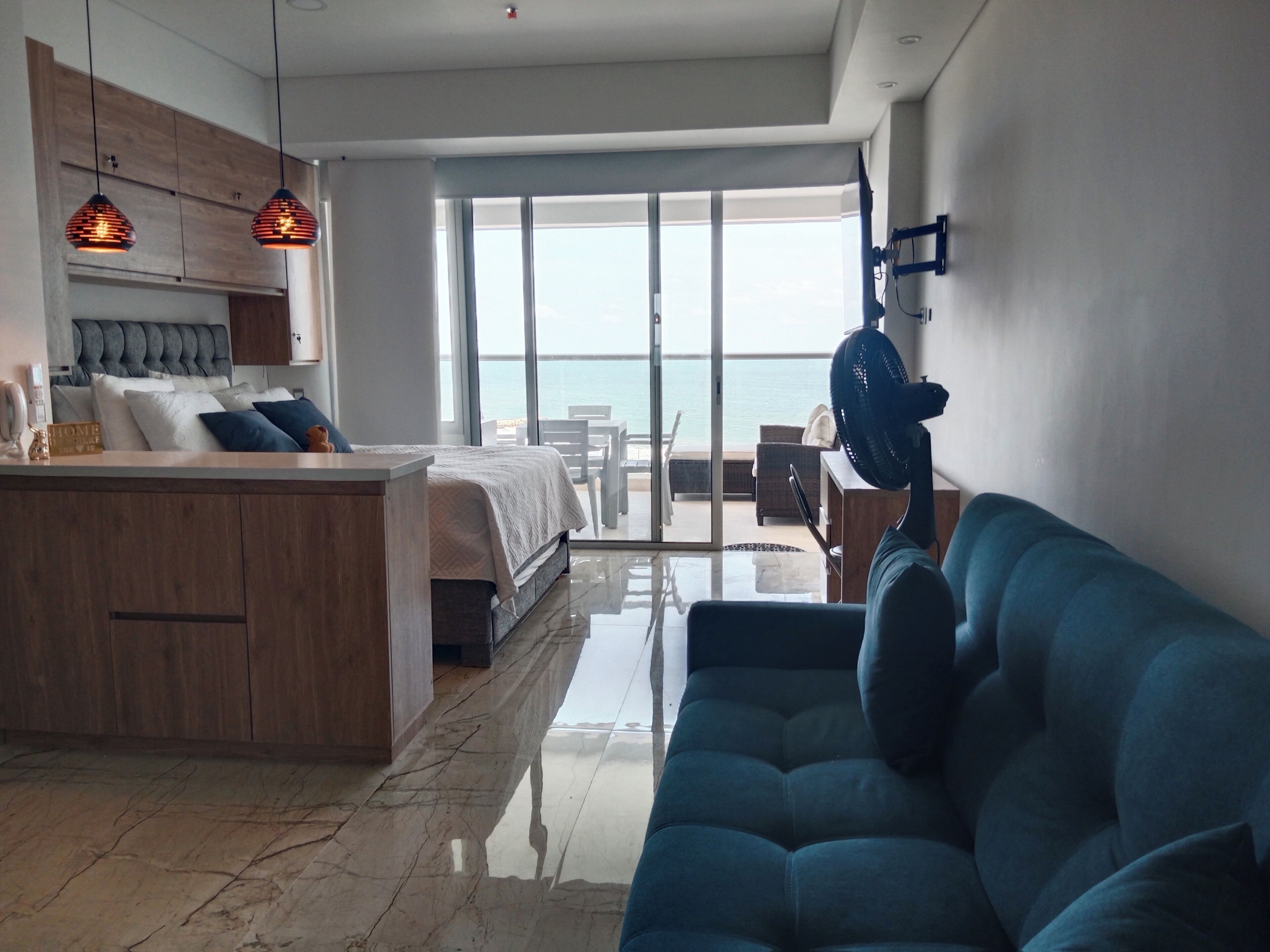A modern hotel room with a bed, a blue sofa, a kitchen area, and a balcony with a view of the sea.