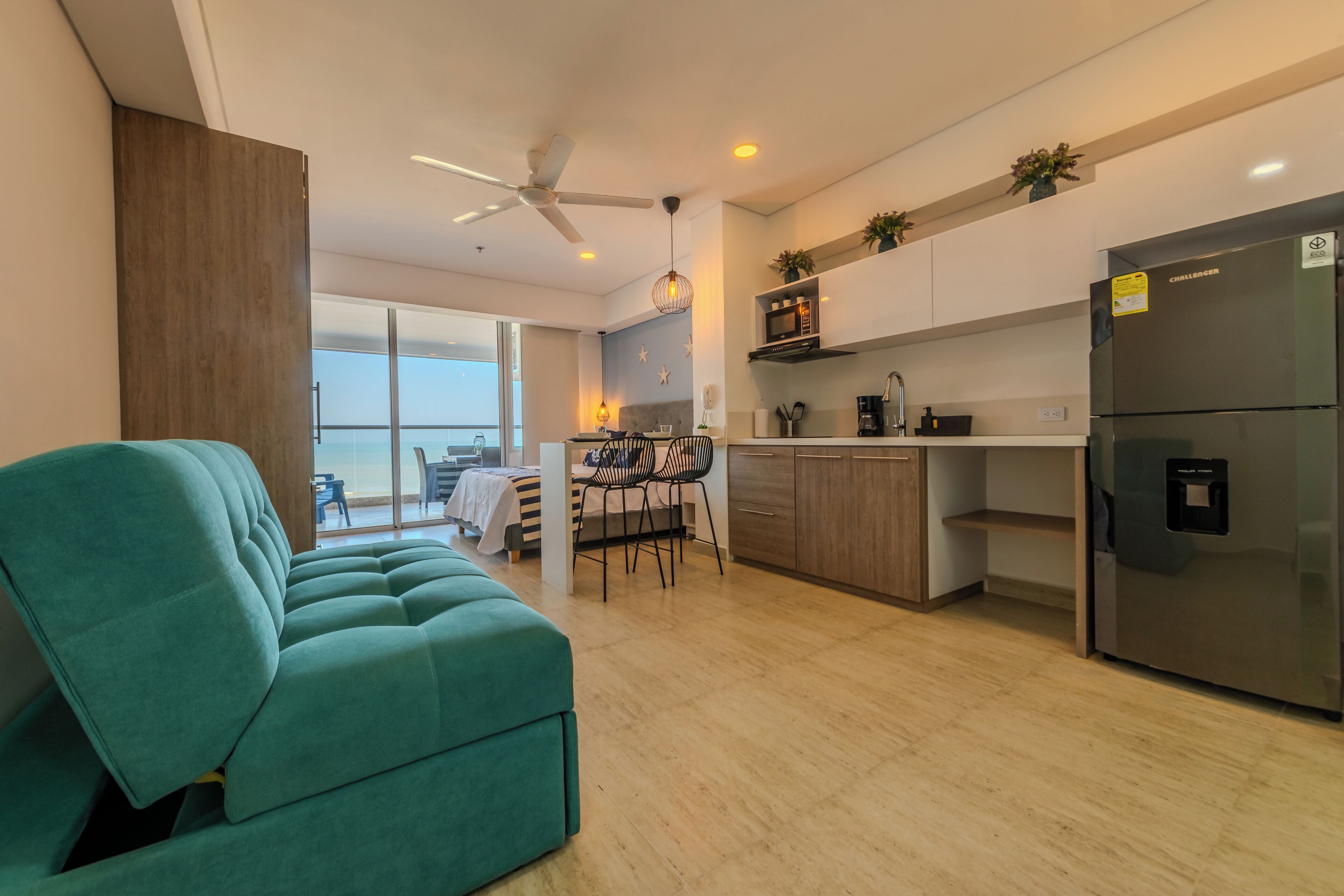 Ein modernes Wohnzimmer mit einem türkisfarbenen Sofa, einem Essbereich mit Tisch und Stühlen, einer Küche mit Kühlschrank und Schränken sowie einem Deckenventilator.