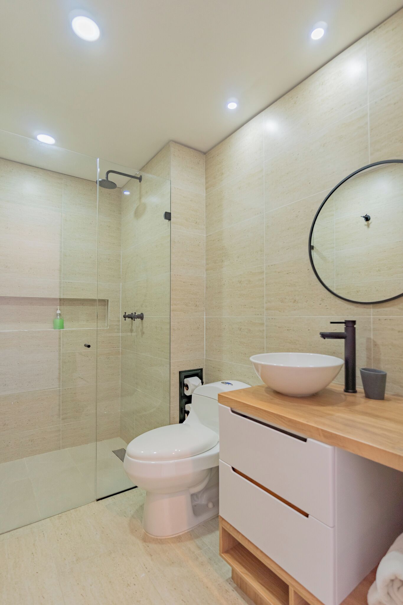 Un baño moderno con cabina de ducha de vidrio, inodoro blanco, tocador de madera con un espejo redondo y pared revestida de baldosas color beige.