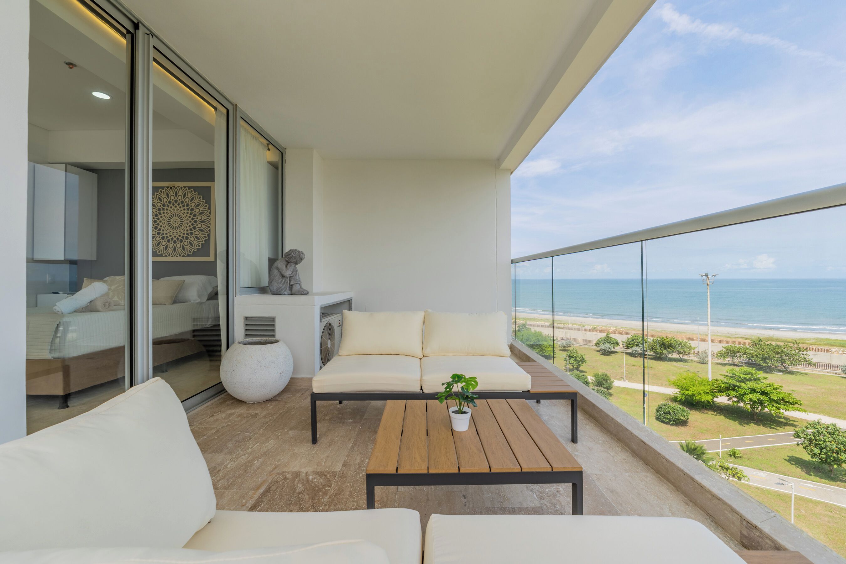 A balcony with a sofa, a small table, and a potted plant. A bed with white bedding is visible through a glass door. The balcony overlooks a beach and the ocean.