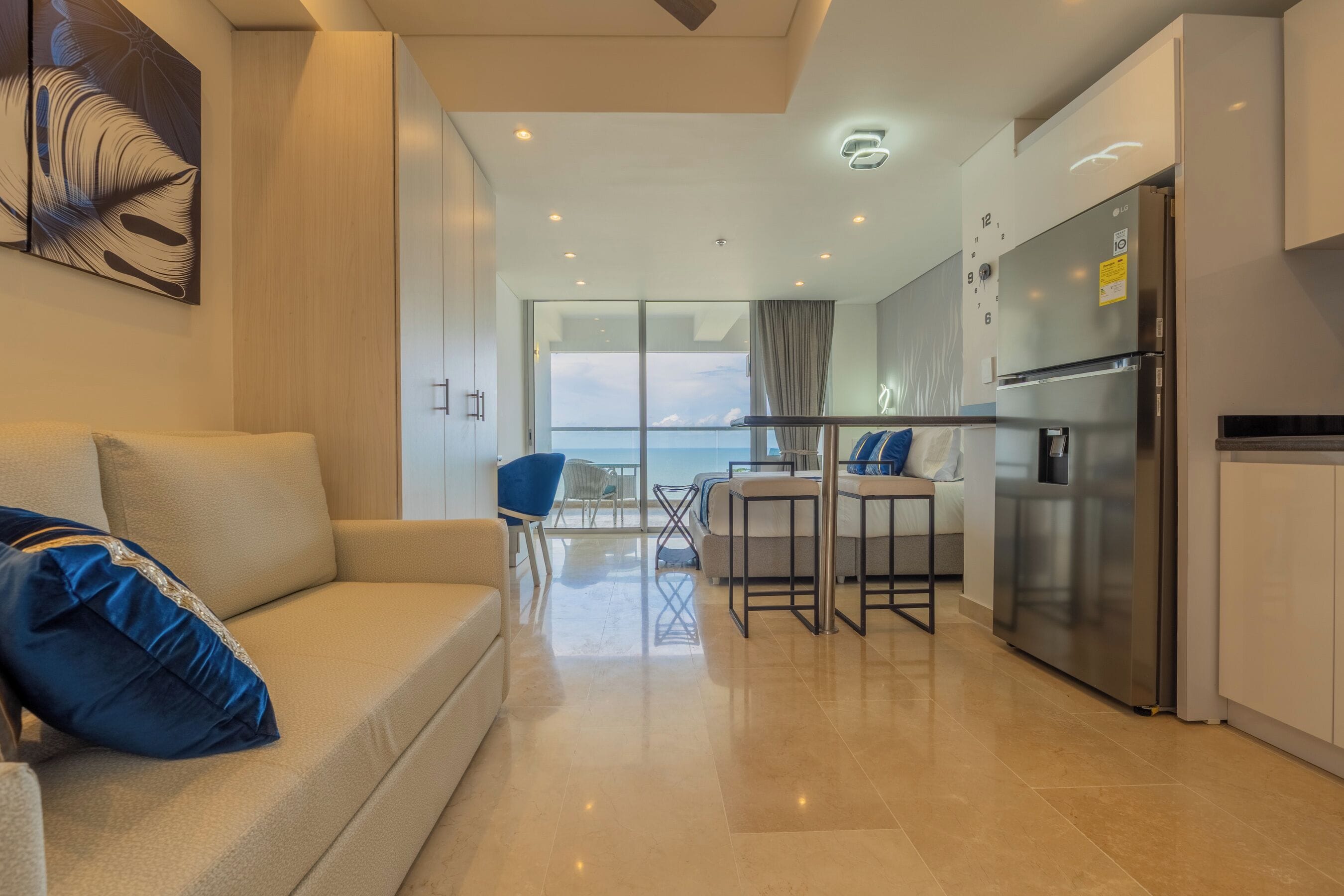 A modern living room with a sofa, a dining area with a table and chairs, a kitchen with a refrigerator, and a large window offering a view of the sky.