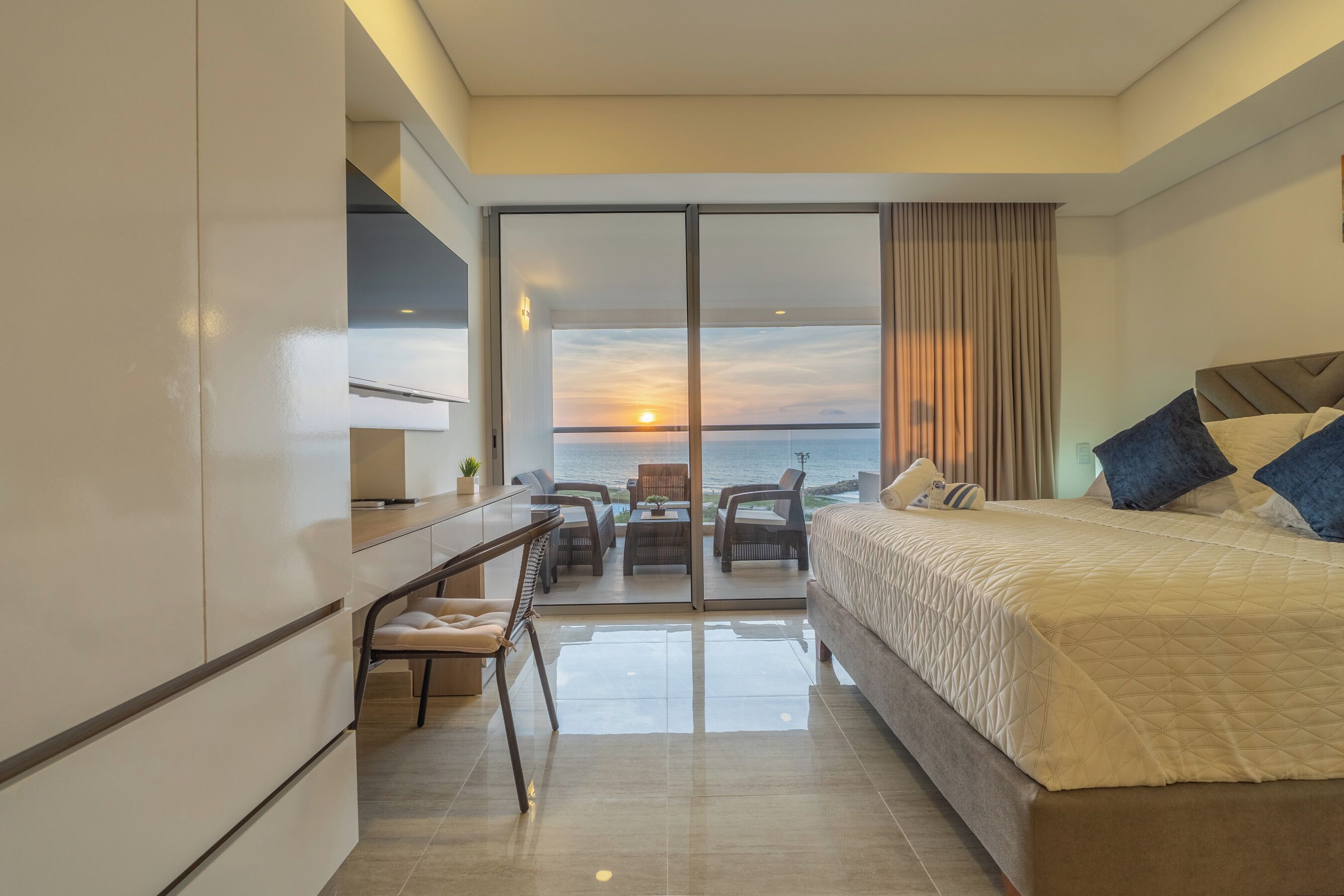 A modern hotel room with a large bed, a desk, and a view of the sunset over the sea.