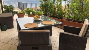 A patio with a table set for dining, surrounded by wicker chairs, potted plants, and a view of the sea and mountains.