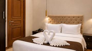 A hotel room with a large bed, a wooden door, a nightstand, and a wall-mounted light fixture.