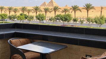 A terrace with a table and chairs, overlooking a street with palm trees and a view of the pyramids.