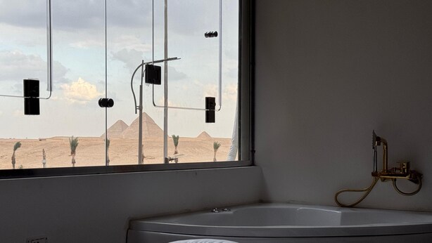 A bedroom with a large window, a bathtub, and a bed with white bedding. - New White Pyramids Inn (Giza)