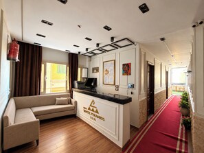 Un hall d’hôtel avec un comptoir d’accueil, un canapé, une moquette rouge et une vue sur l’extérieur.