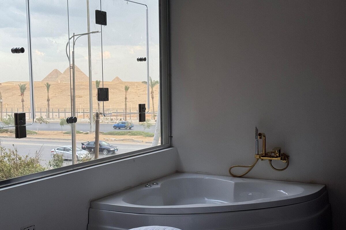 A modern bathroom with a large bathtub, a window with a view of pyramids, and a gold faucet.