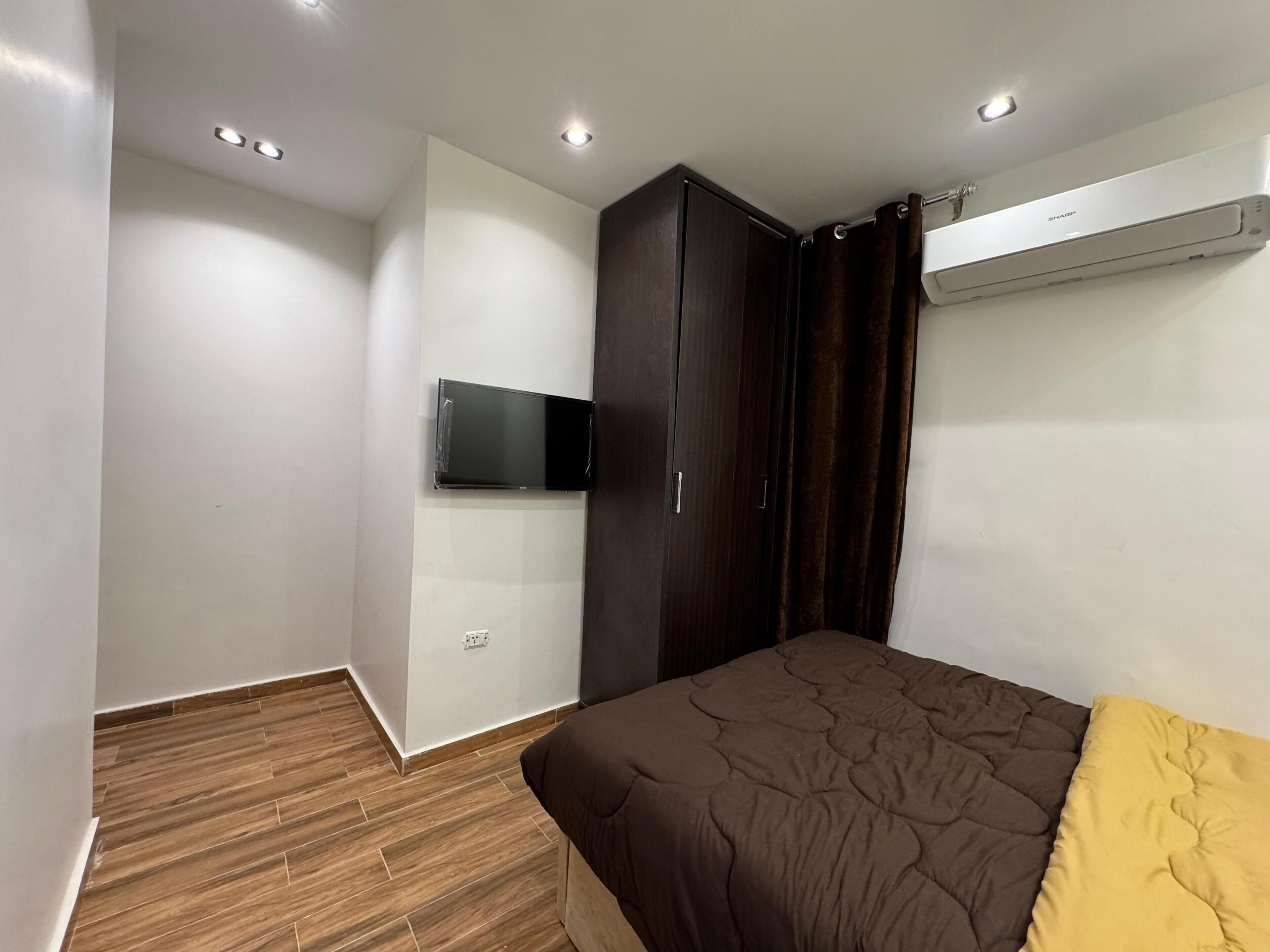 A bedroom with a bed, a wardrobe, a flat-screen TV mounted on the wall, and an air conditioning unit.