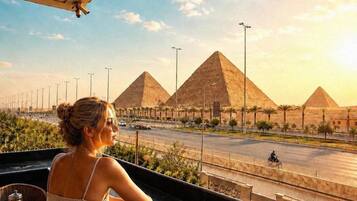 A person seated at a table with a tray containing food and a drink, overlooking a street with palm trees and a view of the pyramids.