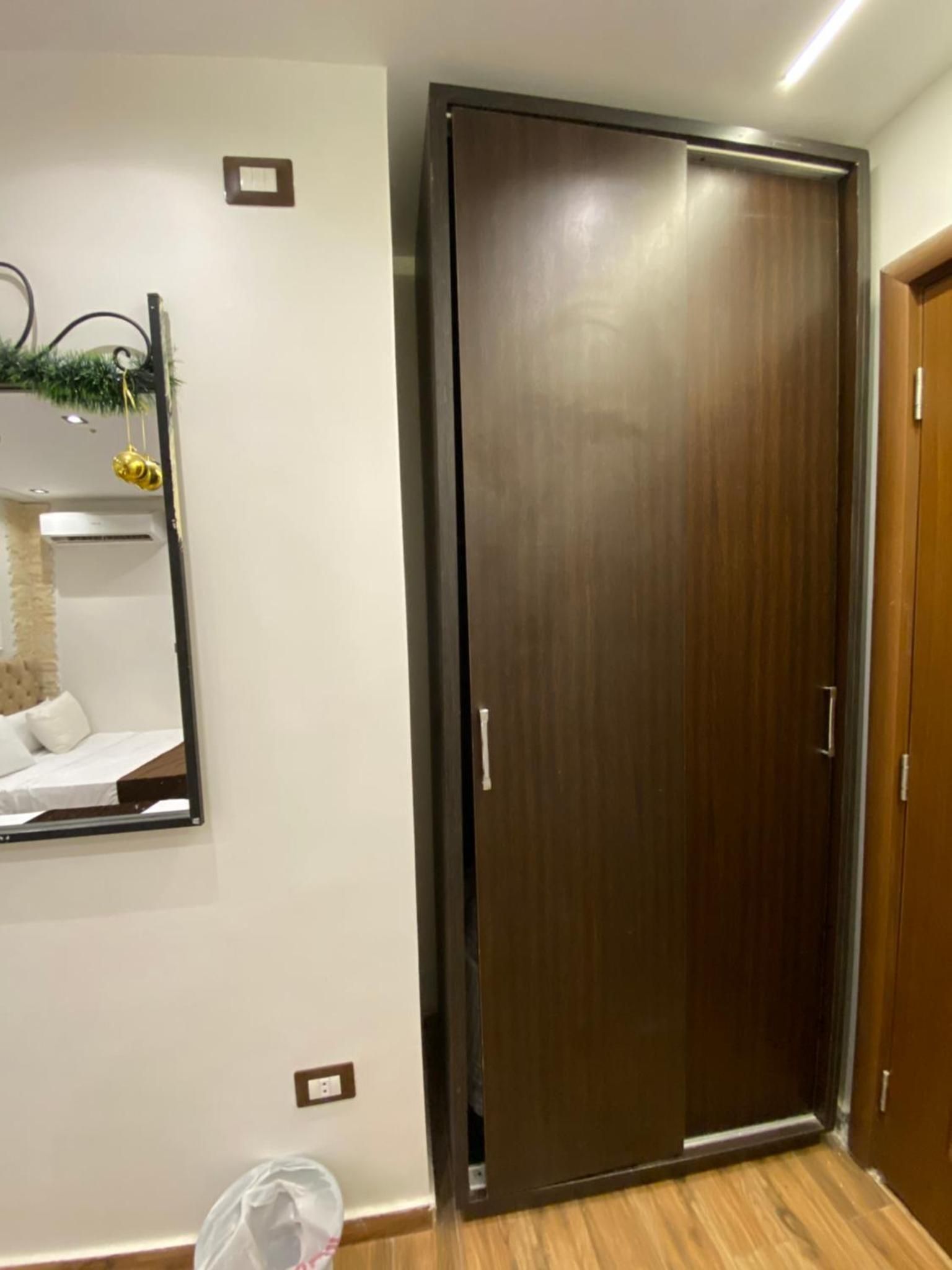 A room with a wooden door, a mirror on the wall, and a small trash bin.