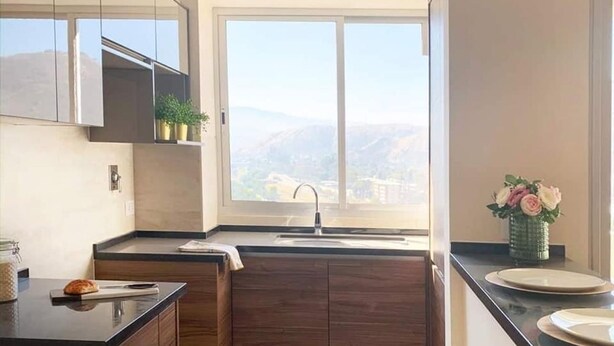 A modern kitchen with dark wooden cabinets, a large window with a city view, and a small vase of flowers on the counter. - 1-bedroom apartment in Cochabamba (Cochabamba)