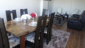 A dining area with a marble table, dark chairs, and a small vase with flowers. There is a sofa and a TV in the background. - 1-bedroom apartment in Cochabamba (Cochabamba)