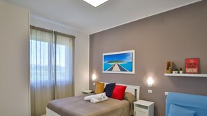 A modern hotel room with a bed, a desk, a chair, a painting, and a shelf with a sign.