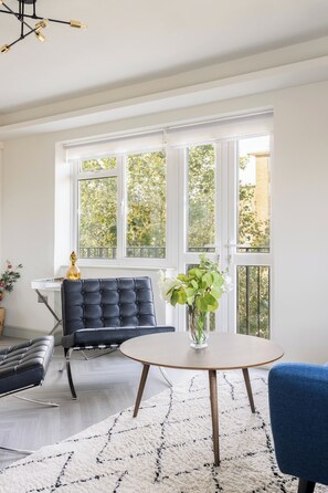 A modern living room with a black sofa, a round coffee table, and a blue chair. There is a large window with white frames and a view of greenery outside. - Lux Regents Park 2 bed 2 bath flat with AC (London)