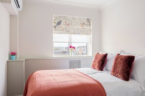 A bedroom with a bed, a window with a view of buildings, a radiator, and a small plant on the windowsill. - Lux Regents Park 2 bed 2 bath flat with AC (London)