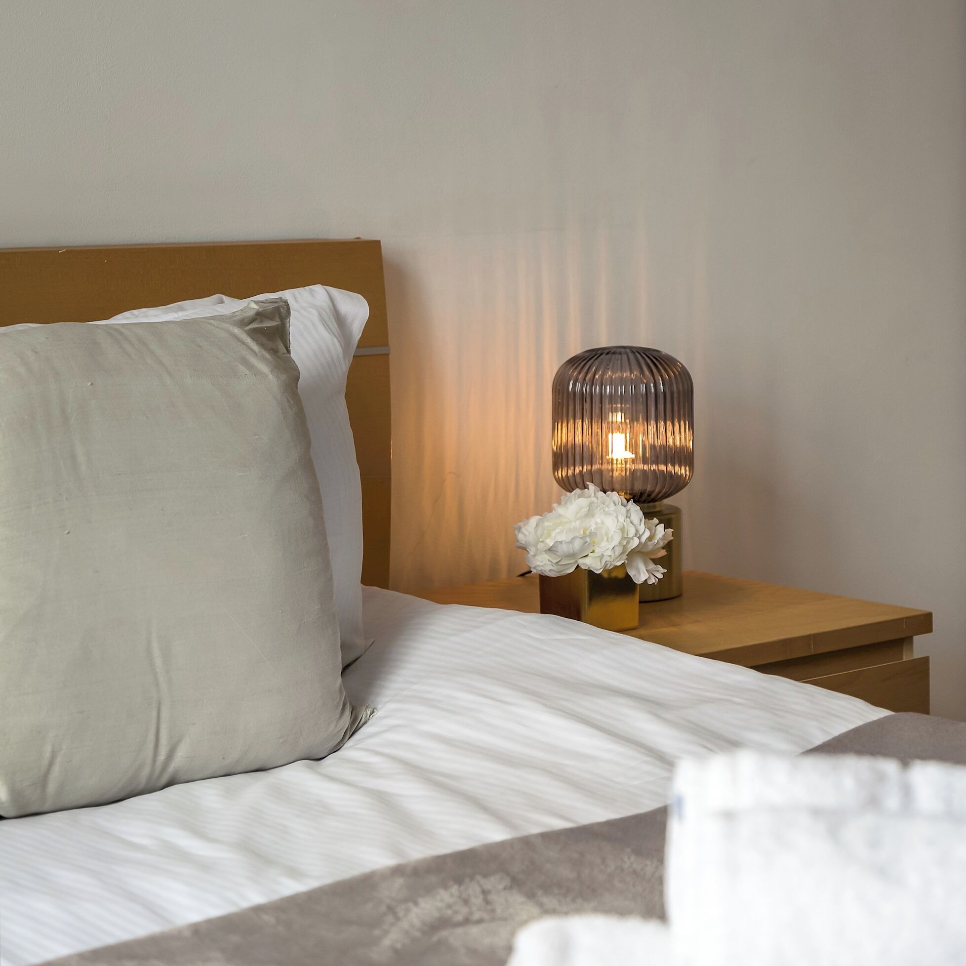 Un lit bien fait, avec du linge de lit blanc et un grand oreiller, une table de chevet agrémentée d’une lampe décorative et d’un vase de fleurs, et une tête de lit en bois.