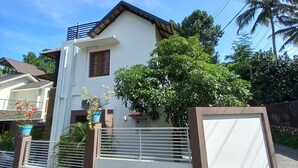 A two-story house with a white exterior and a brown roof. There is a large tree in front of the house, and a metal gate with a white panel.
