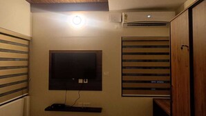 A small room with a flat-screen TV mounted on the wall, a ceiling fan, and a window with horizontal blinds.