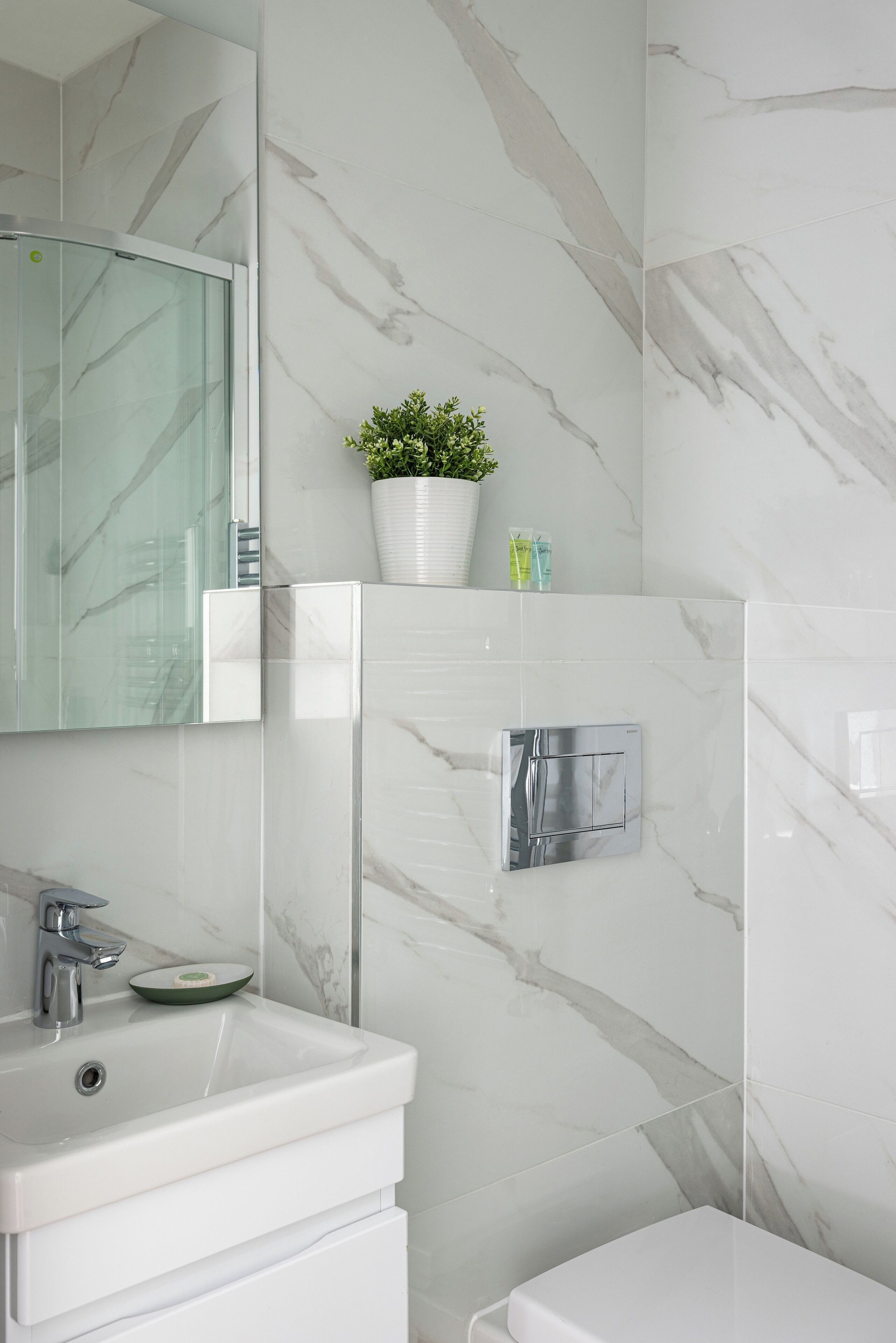 A bathroom with a white sink, a mirror, a potted plant, and a shower control panel.