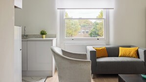 Una cocina moderna con armarios blancos, un sofá gris con cojines amarillos, y una ventana con vista a árboles y edificios.
