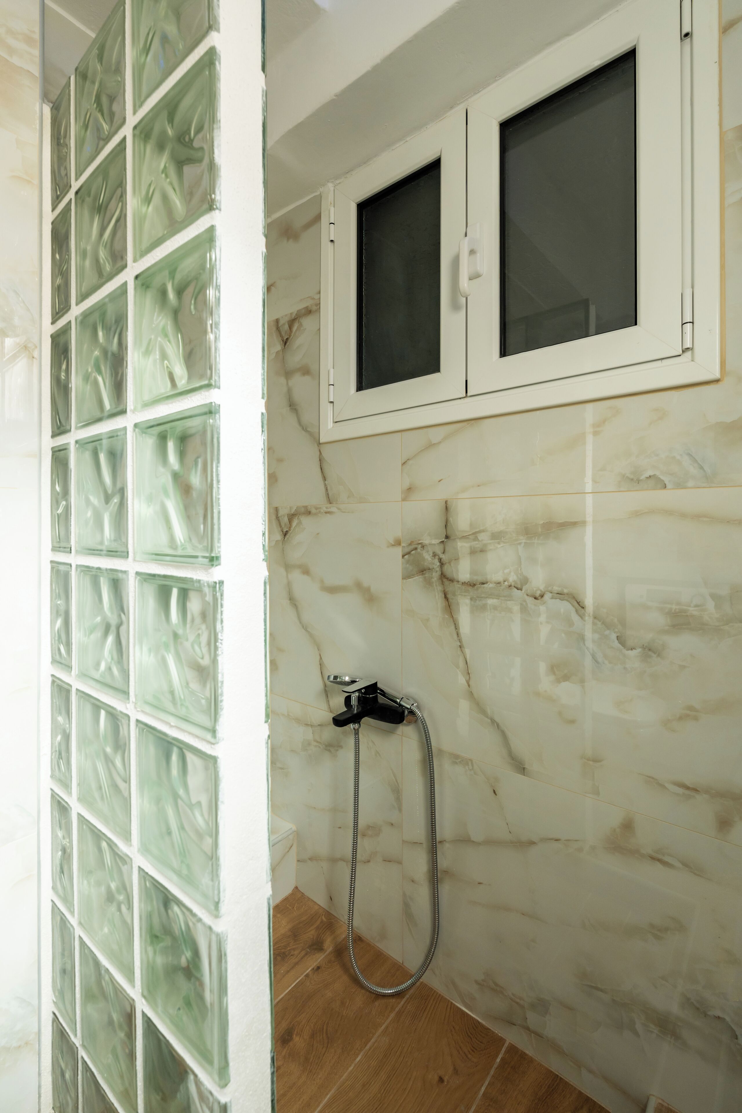 A bathroom with a glass block wall, a shower with a handheld hose, and a marble-tiled wall.