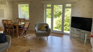 Uma sala de estar com piso de madeira, uma poltrona cinza, uma mesa de jantar com cadeiras, um sofá, uma TV em um suporte de vidro e janelas amplas com vista para vegetação.